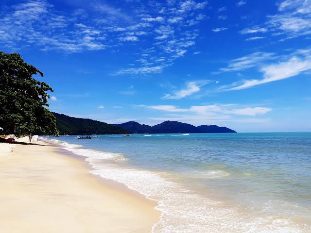 Tropical beach with white sand, blue ocean, lush green trees, distant islands and clear sky.