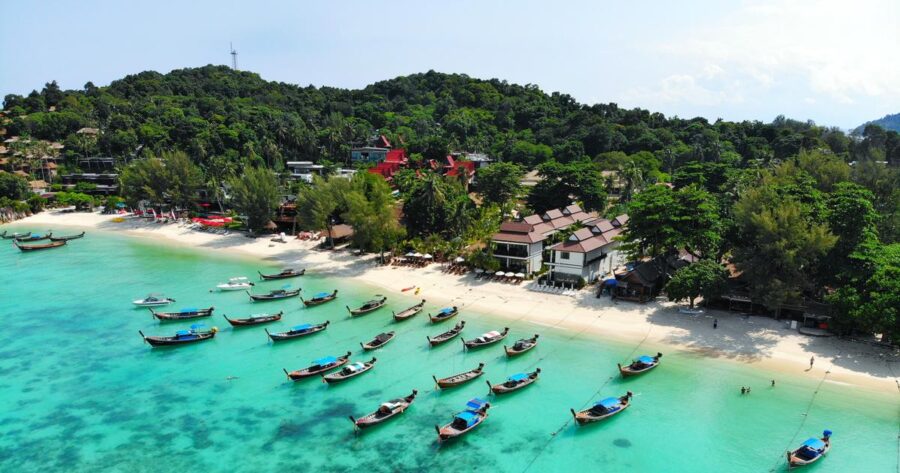 Aerial view of tropical beach with longtail boats, turquoise water, palm trees and coastal village.