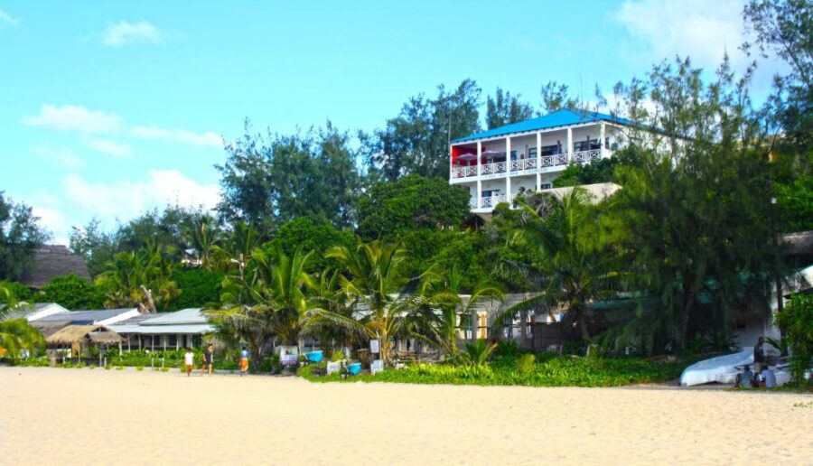 Tropical beachfront guesthouse behind sandy beach with palm trees, rustic huts and lush green hillside.