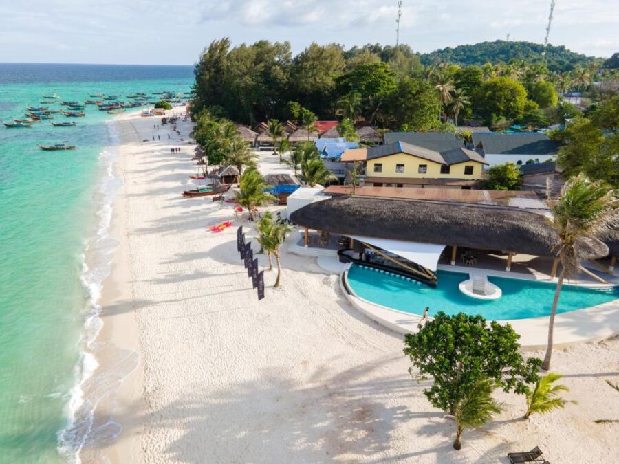 Luxury tropical beachfront resort with infinity pool, white sand beach, long-tail boats, palm trees, and villas.