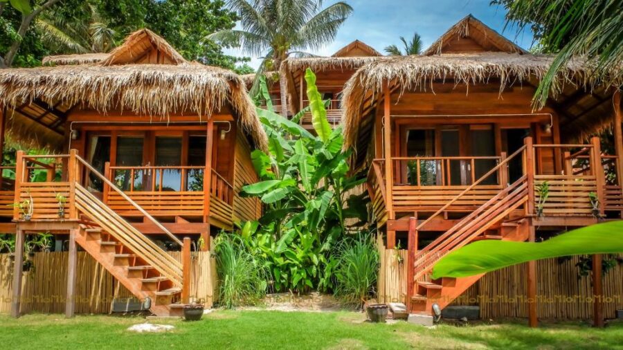 Tropical wooden bungalows with thatched roofs and lush green garden at beach resort.