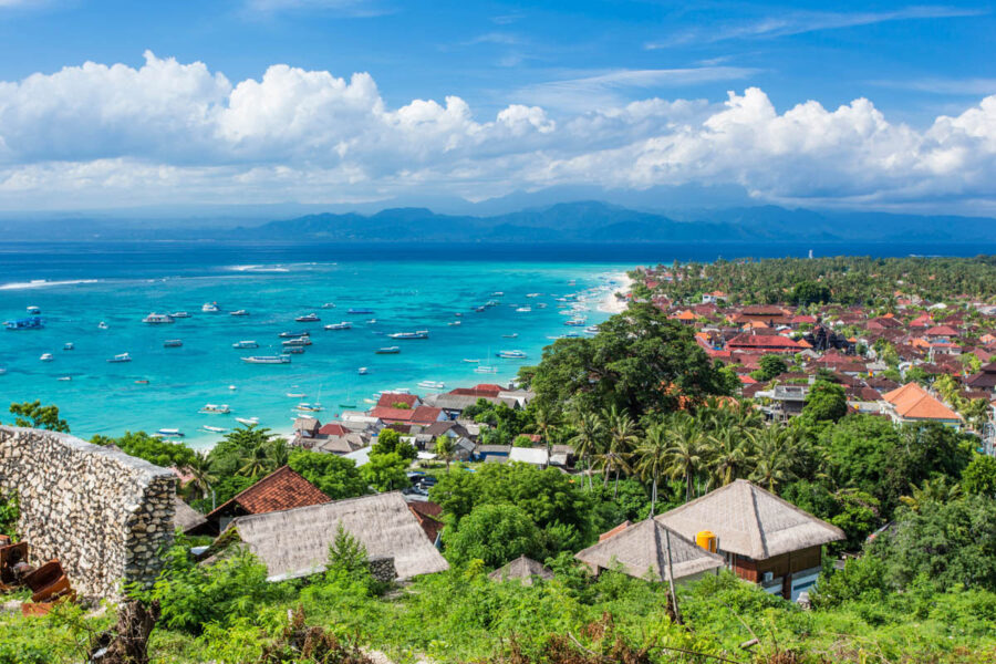 Tropical Indonesian coastal village with turquoise bay, fishing boats, sandy beach, lush greenery, and mountains.