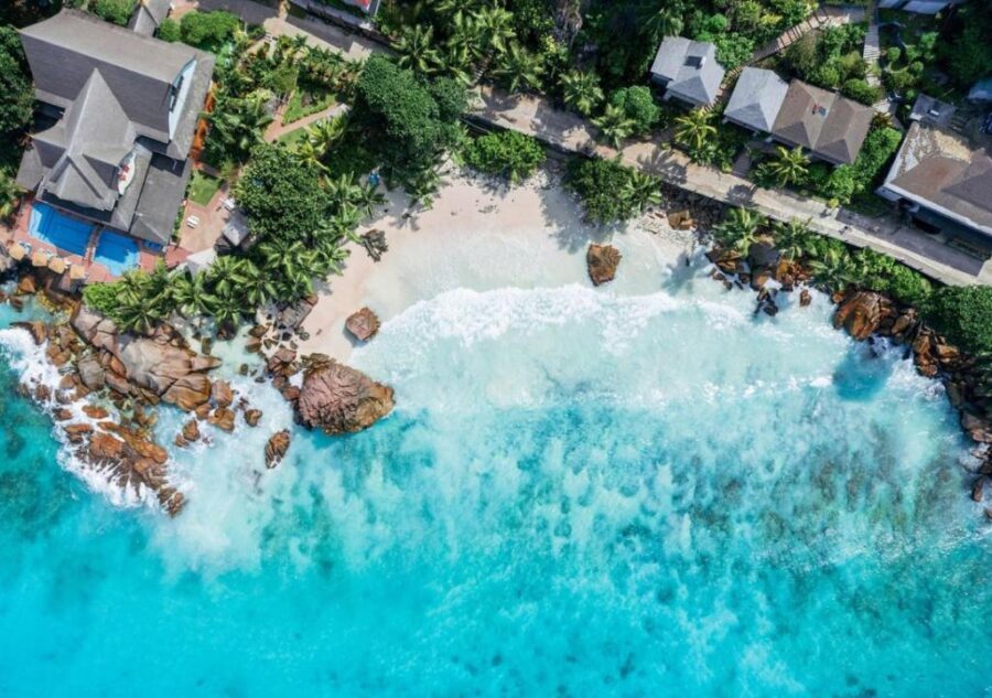 Aerial view of tropical beach cove, luxury villas, palm trees, rocks, turquoise ocean coastline.