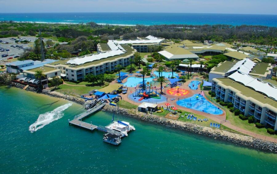 Aerial view of tropical family resort waterpark with lagoon, pools, oceanfront hotel and catamaran.