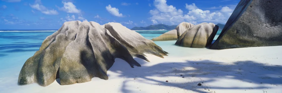 Tropical granite boulders on white sand beach with turquoise ocean and palm tree shadows.