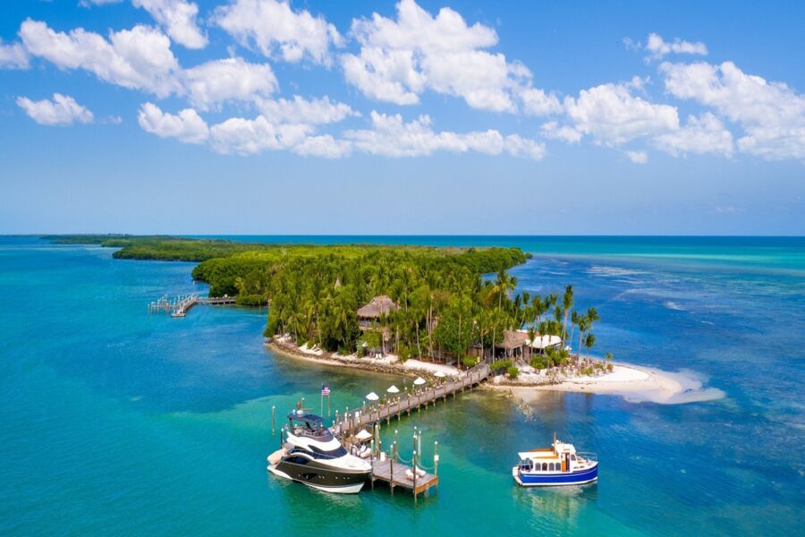 Aerial view of lush private island with turquoise waters, sandy beach, yacht, and palm trees.