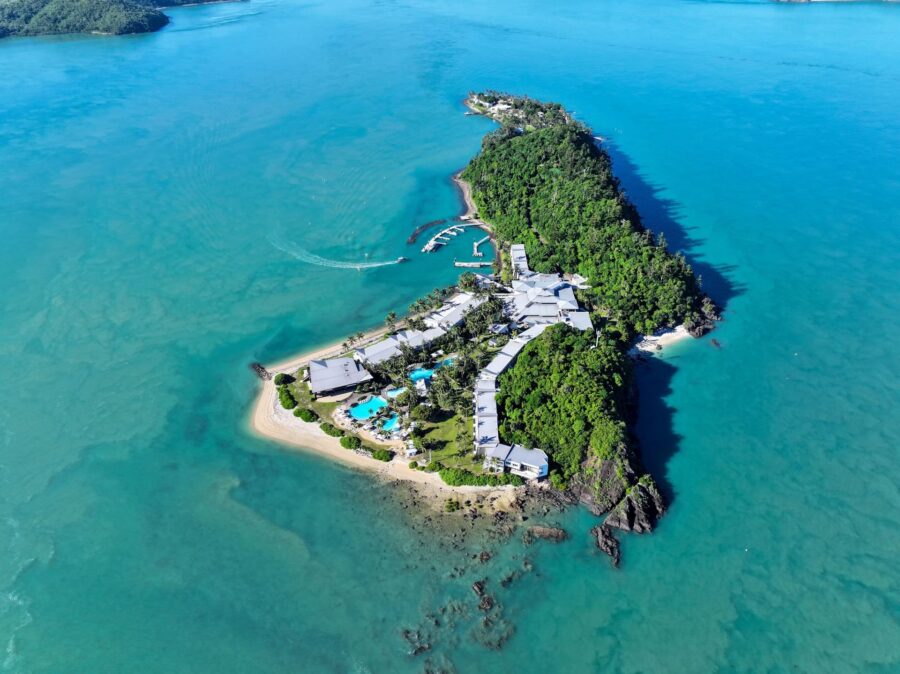 Aerial view of tropical island resort with beaches, palm trees, turquoise ocean, luxury villas, and marina.