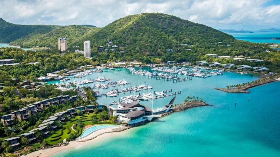 Aerial view of tropical island resort marina with yachts, beach, lush green hills and luxury hotels.