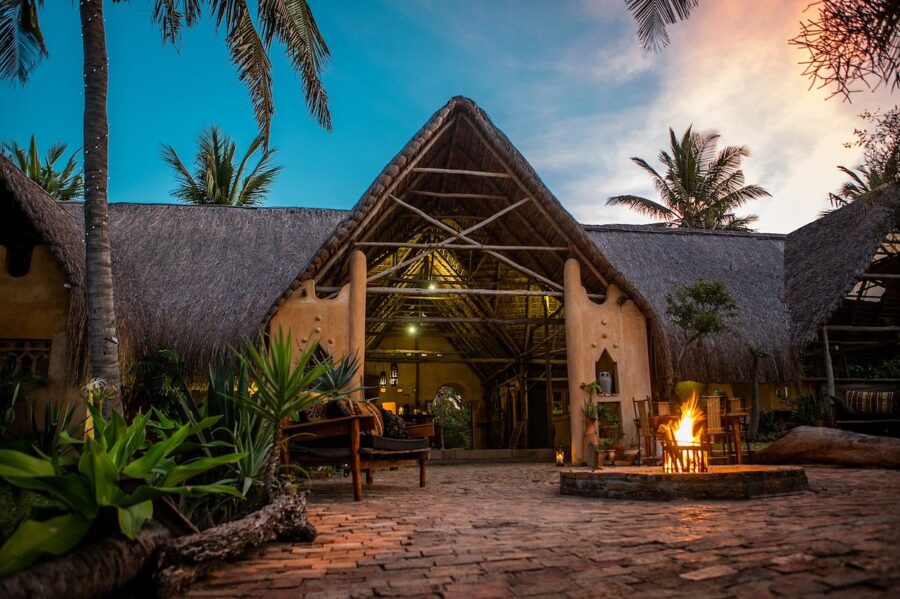 Tropical lodge at dusk with thatched roof, palm trees, glowing fire pit, and outdoor seating.