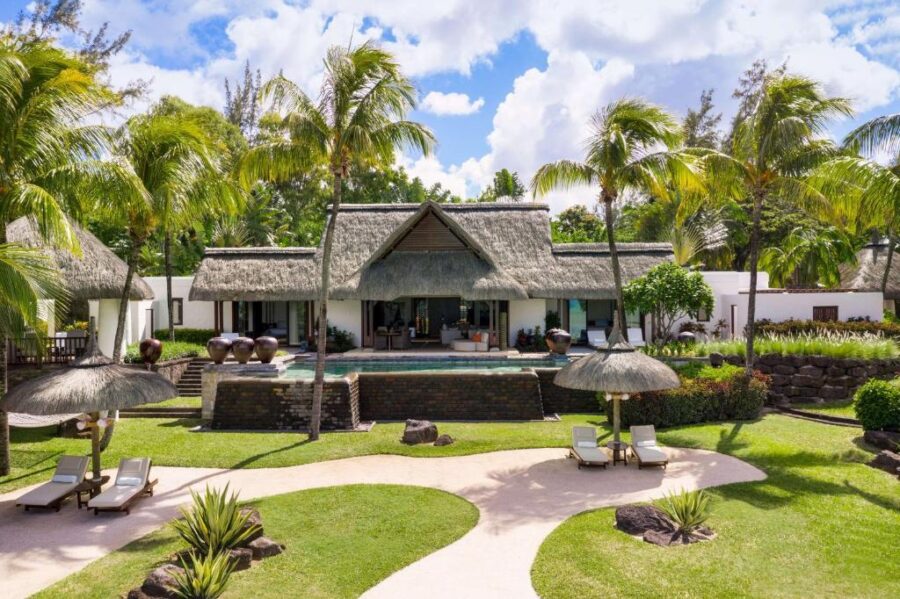 Luxury tropical villa resort with infinity pool, palm trees, thatched roofs, and landscaped beachfront garden.
