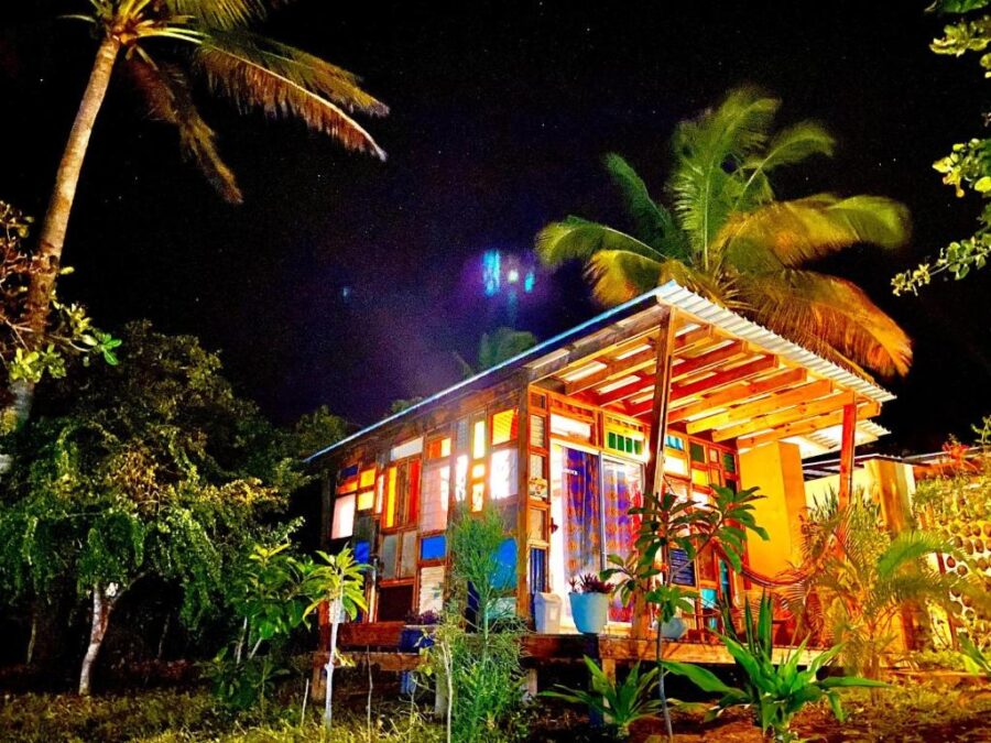 Colorful tropical cabin glowing at night surrounded by palm trees, lush plants and starry sky.