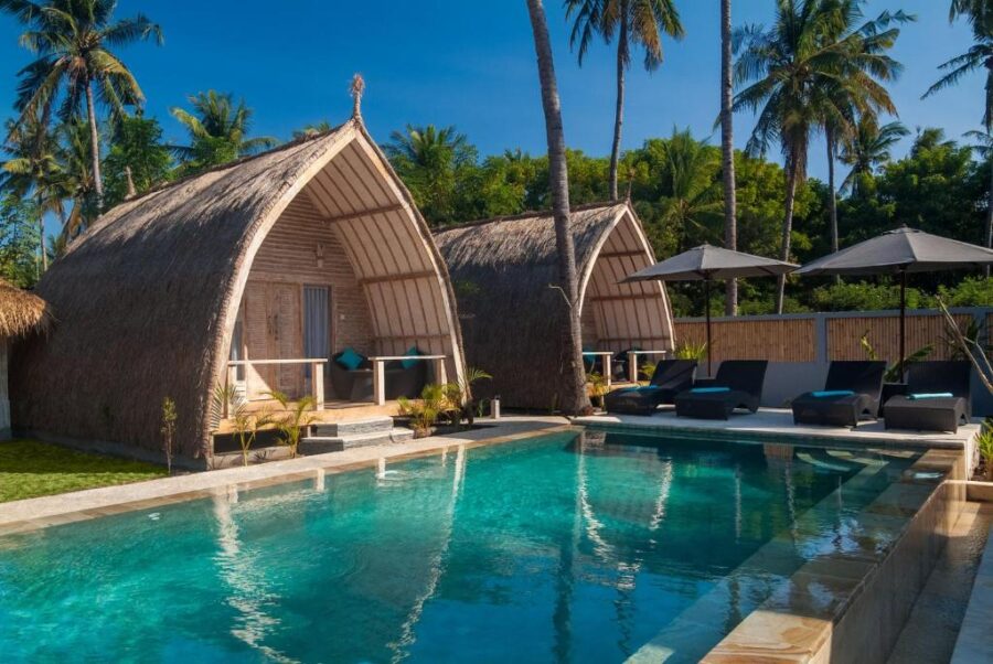 Tropical island resort bungalows with thatched roofs beside a turquoise swimming pool and palm trees.