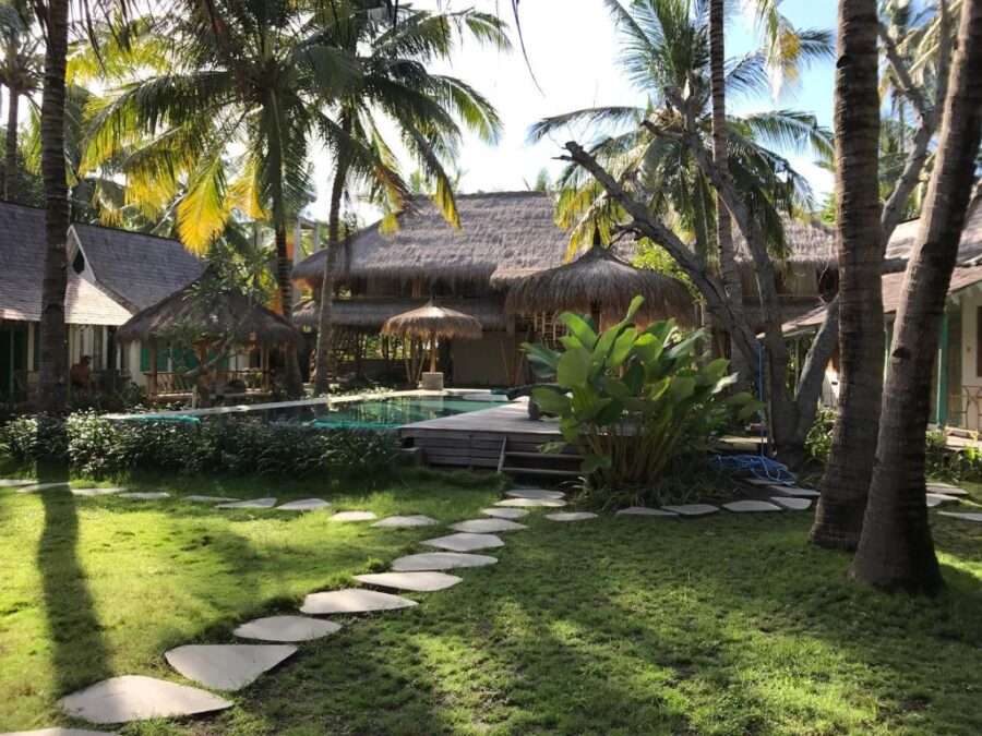 Tropical resort garden with swimming pool, thatched villas and coconut palm trees at serene island retreat.