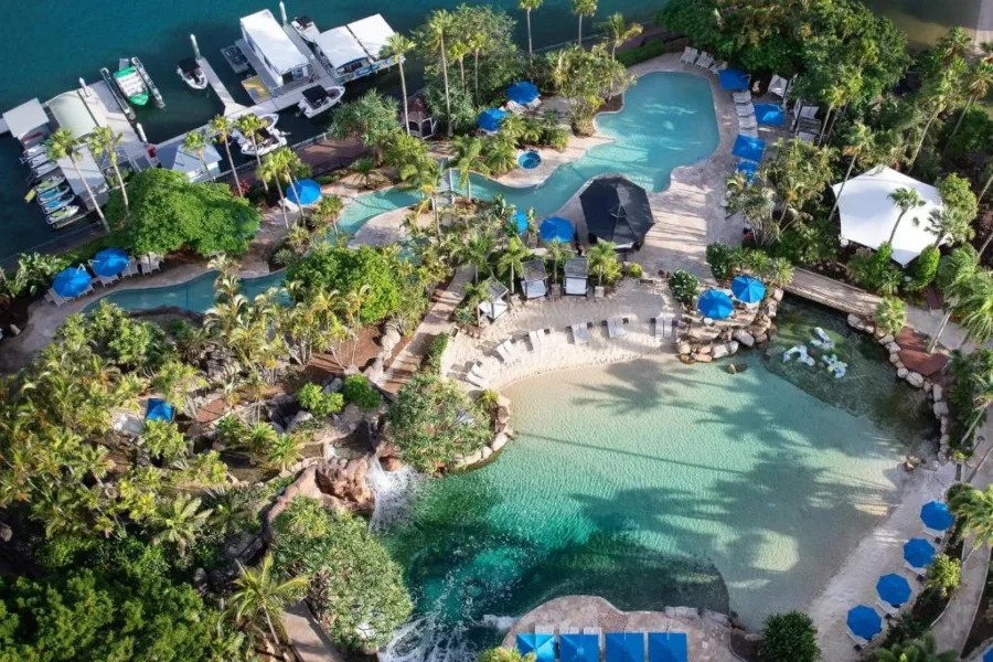 Aerial view of tropical resort lagoon pools with palm trees, sandy beach entry, and marina.