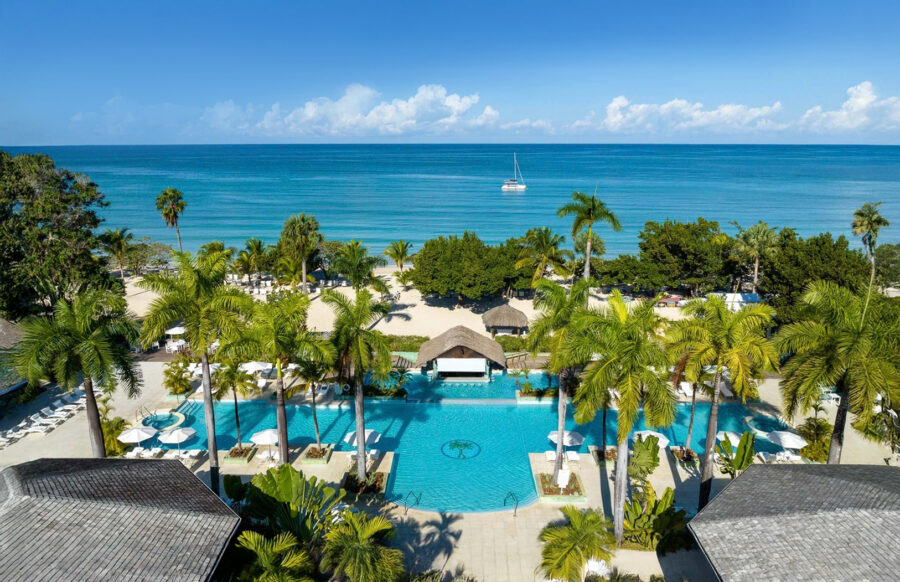 Tropical seaside resort with pool, beach, palm trees, sailboat, and azure sky.