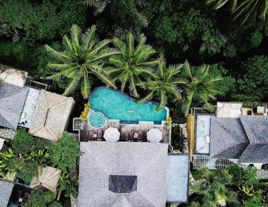 Aerial view of tropical Ubud resort with freeform pool, palm trees, jungle surroundings, and villas.
