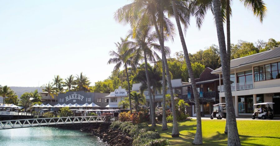Tropical waterfront bakery and fish and chips restaurant with palm trees, promenade, and resort buildings.