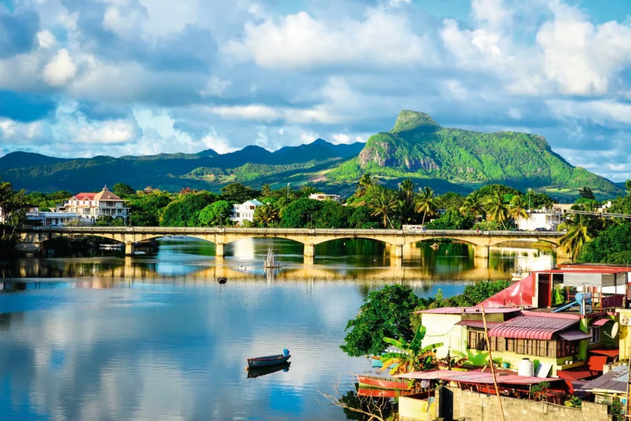 Tropical riverside town with mountain backdrop, arched bridge, colorful houses and calm reflective water.