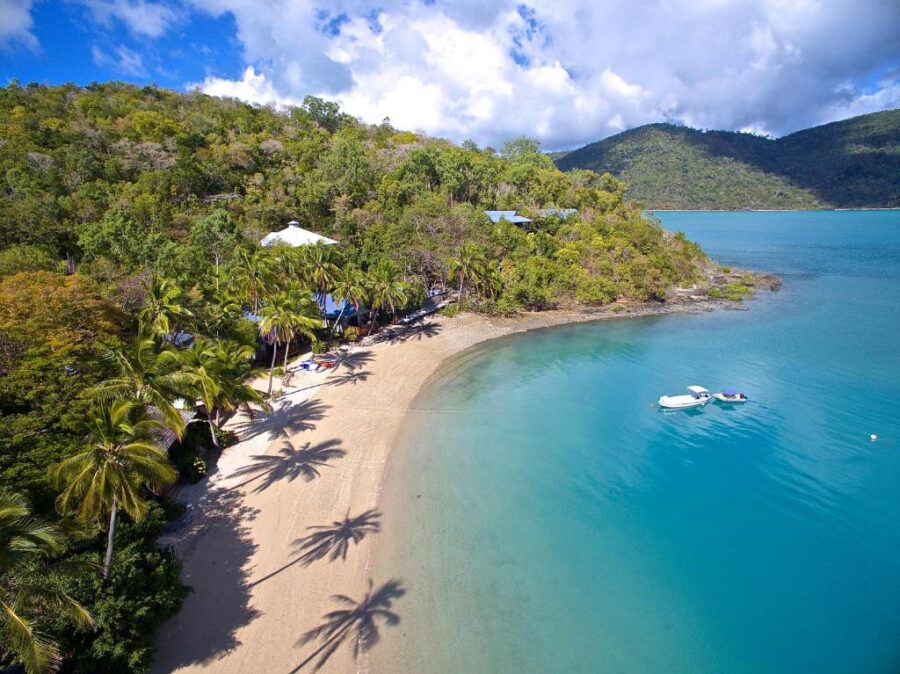 Secluded tropical beach resort with palm trees, turquoise bay, oceanfront villas and anchored boats.