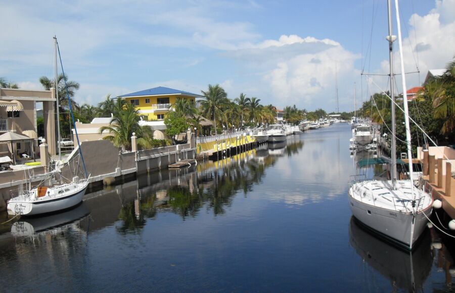 Serene tropical canal with palm trees, sailboats, and colorful waterfront homes under a blue sky.