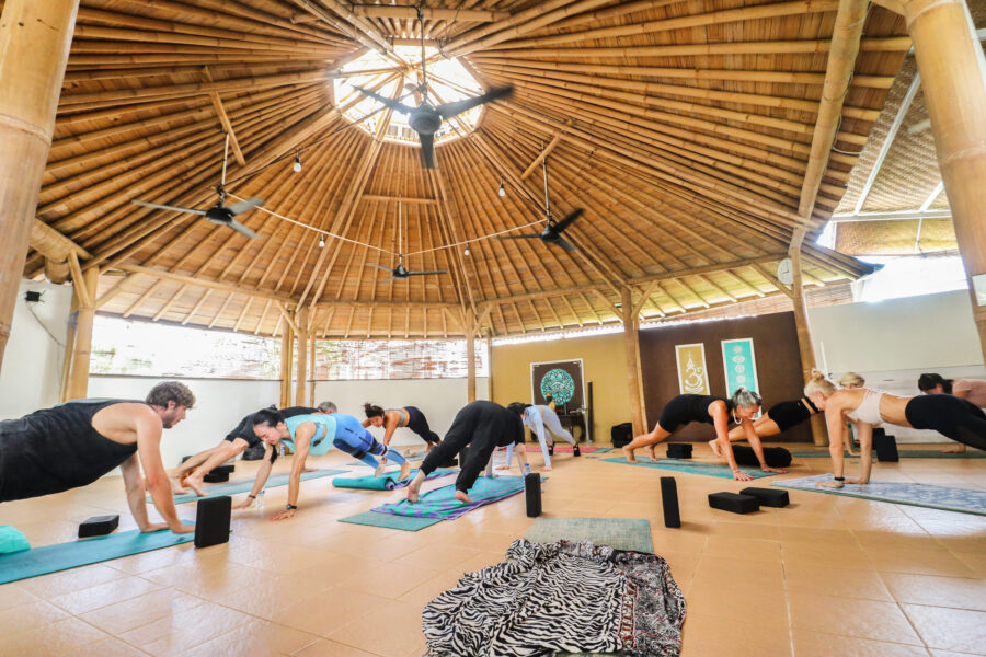 Group yoga class in tropical bamboo pavilion, people holding plank pose on colorful mats.