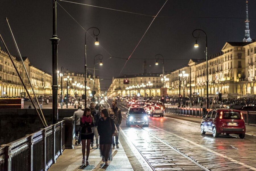 Night scene of bustling Turin boulevard with street lamps, pedestrians, and historical architecture.