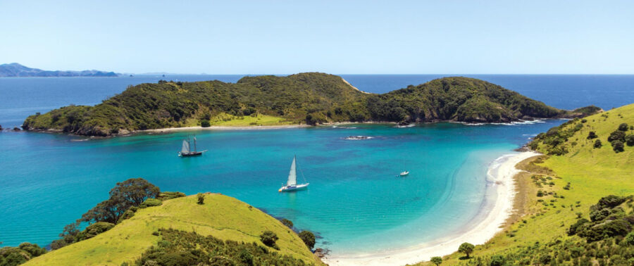 Turquoise New Zealand bay with sailboats, white sand beach, and lush green hills.