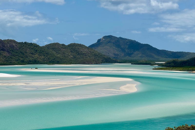 Aerial view of turquoise coastal inlet with white sandbars, tropical hills, and blue sky.