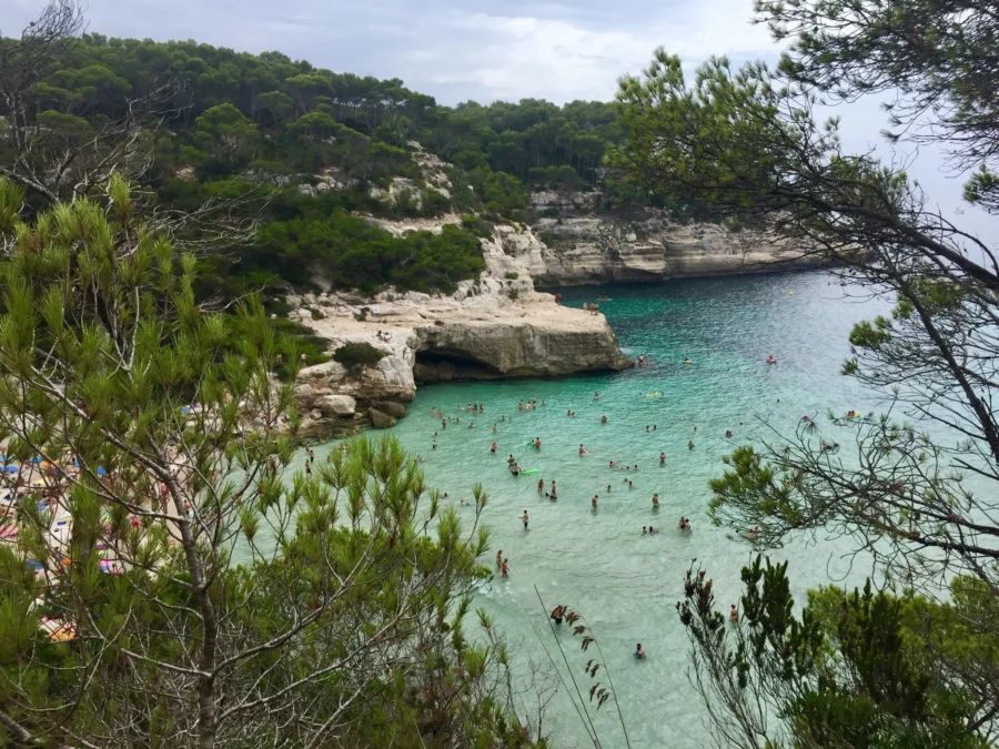 Cala Mitjana turquoise cove with sandy beach, limestone cliffs, pine trees, swimmers.