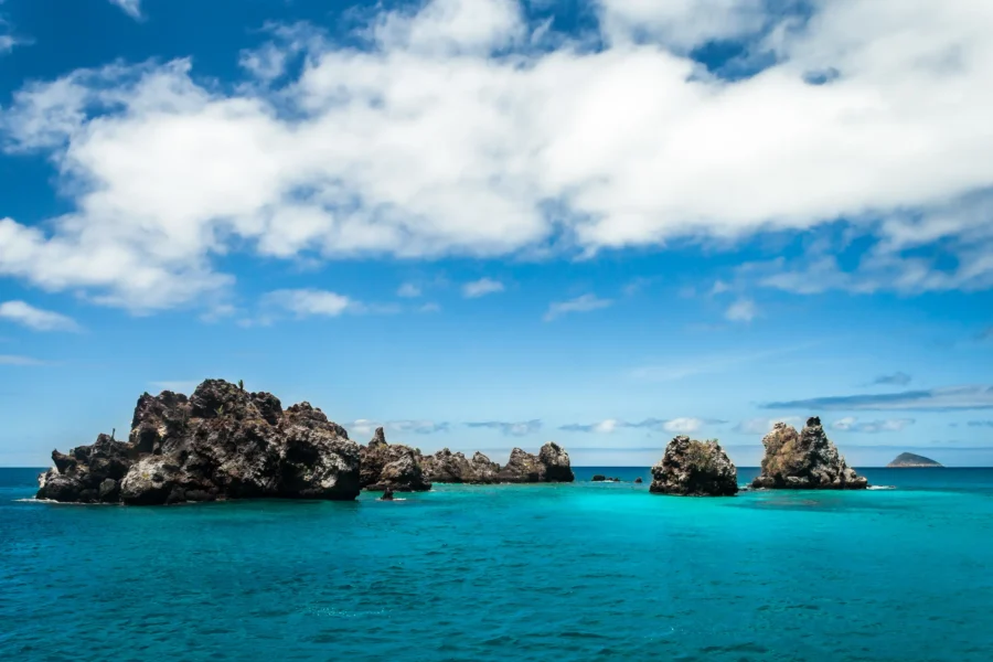 Rocky coastline with turquoise sea and fluffy clouds under a clear blue sky.
