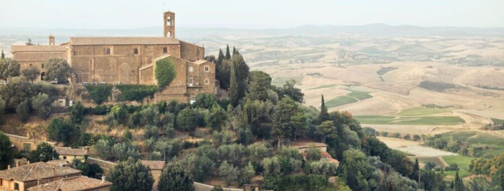 Tuscan countryside with historic monastery, olive and cypress trees, rolling hills, and terracotta roofs.