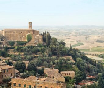 Tuscan countryside with historic monastery, olive and cypress trees, rolling hills, and terracotta roofs.