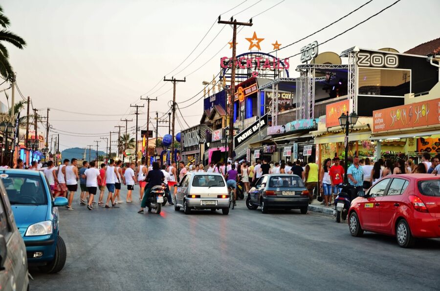 Lively evening street scene with neon lights, parked cars, and a vibrant crowd enjoying nightlife.