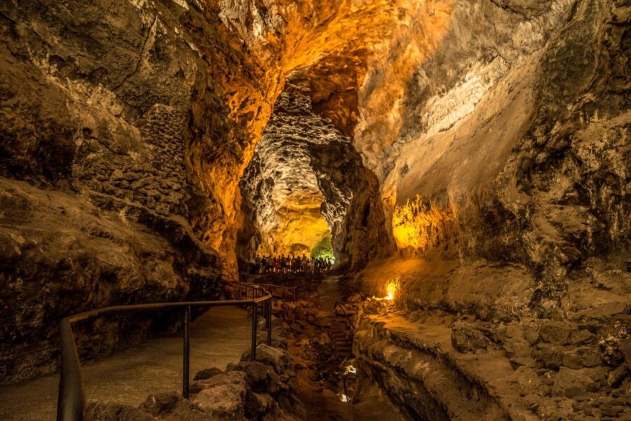 Tourists walking along illuminated lava tube cave walkway with rock formations in Cueva de los Verdes.