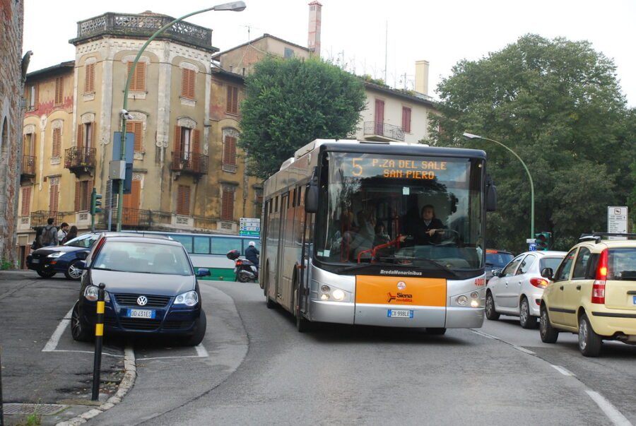 Siena city bus on urban street corner with cars, trees, and historic buildings.