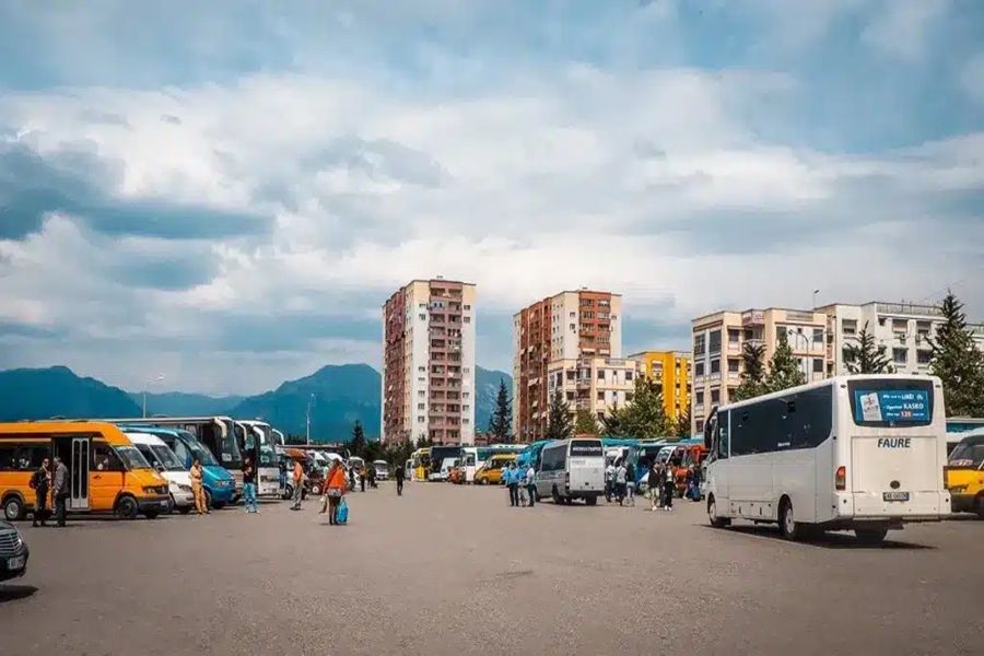 Bustling bus station with colorful buses, travelers, urban backdrop, trees, and distant mountains.