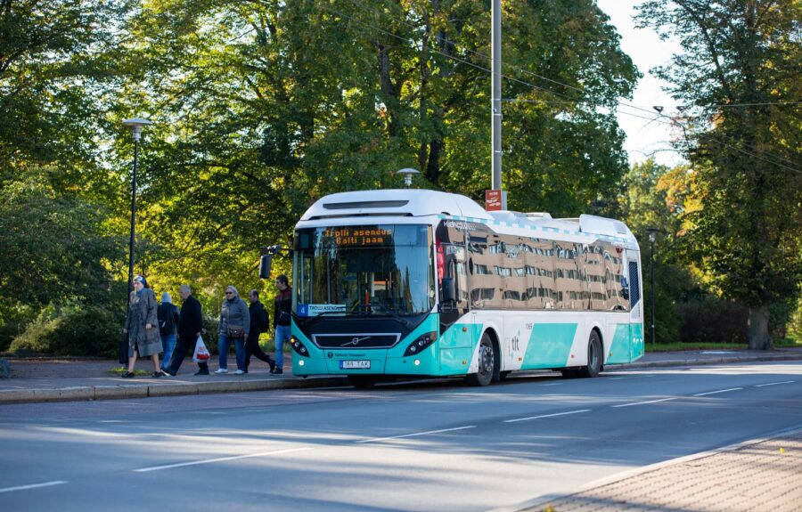 Turquoise Tallinn electric city bus parked by tree-lined sidewalk.