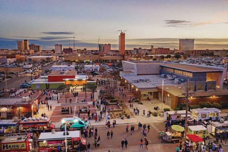 Twilight urban scene with vibrant food trucks, modern architecture, and bustling crowd.