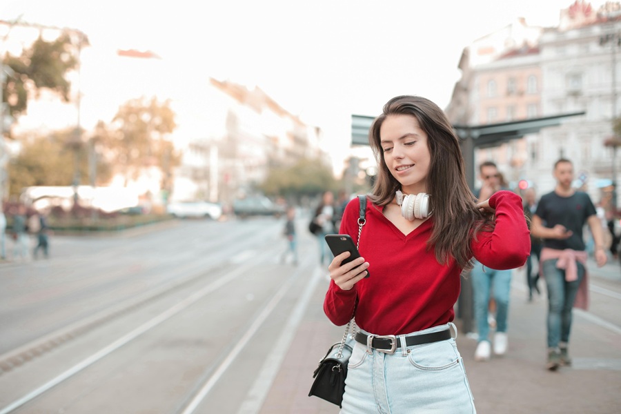 Young woman in vibrant urban setting checks smartphone, wears red sweater and denim shorts.