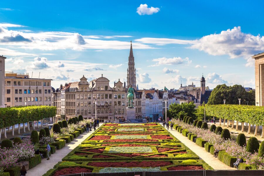 Urban garden in Brussels with colorful flowers, statue, and diverse architecture under blue sky.