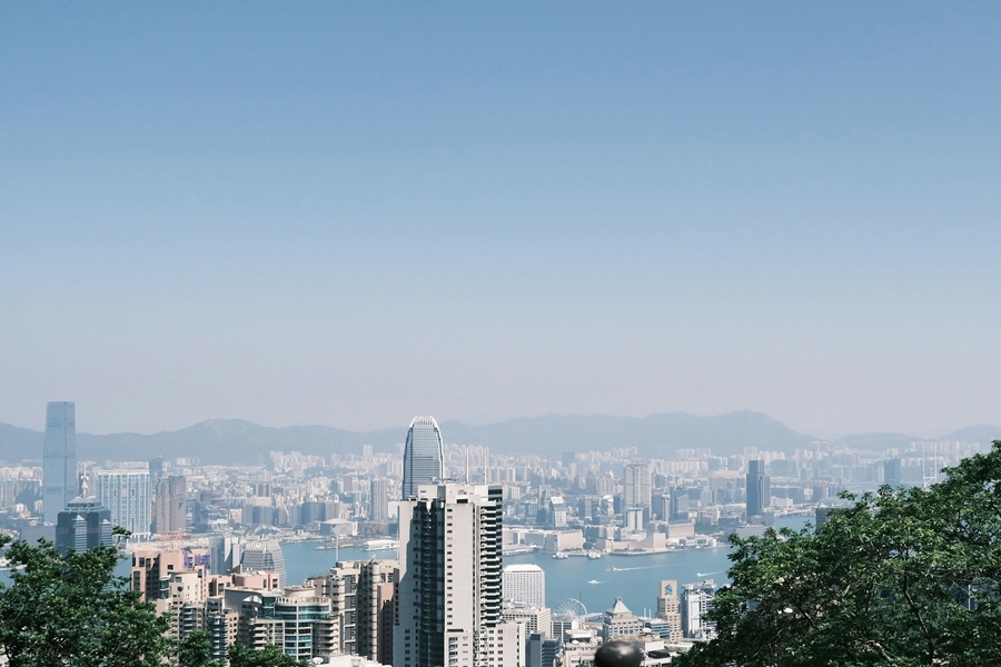 Vibrant urban skyline with high-rises, greenery, water, and mountains under clear blue skies.