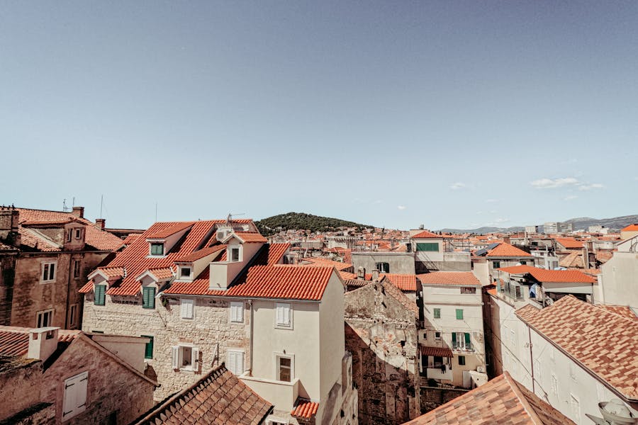 Mediterranean cityscape with terracotta rooftops, stone façades, and lush green hill backdrop.
