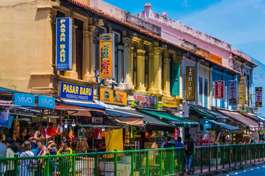 Bustling multicultural market with colorful storefronts and lively shoppers under a clear sky.