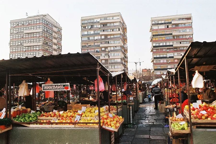 Bustling Belgrade market with colorful produce, Cyrillic signs, urban backdrop, community interaction.