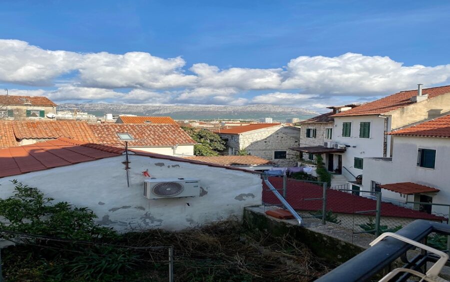 Picturesque Mediterranean neighborhood with terracotta rooftops, blue sky, clouds, and distant mountains.