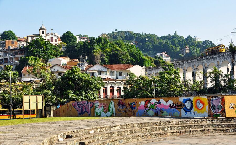 Urban scene with plaza, hills, aqueduct, train, traditional architecture, and vibrant mural.