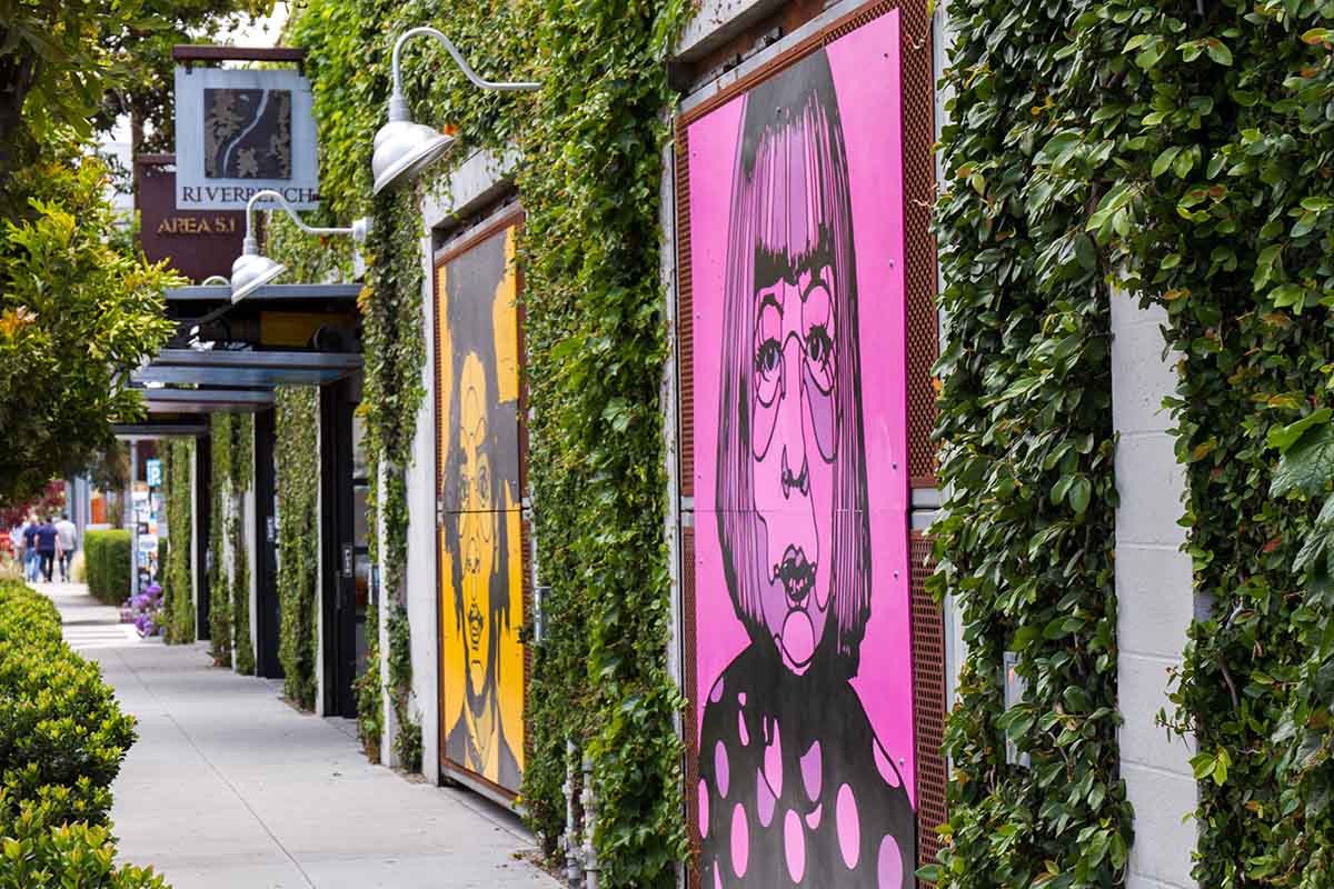 Vibrant urban street with colorful murals, green hedges, and lively community ambiance.