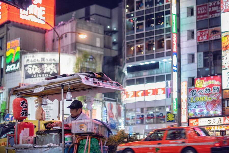 Urban night scene: vibrant neon, food vendor, bustling street, taxis, dynamic cityscape.