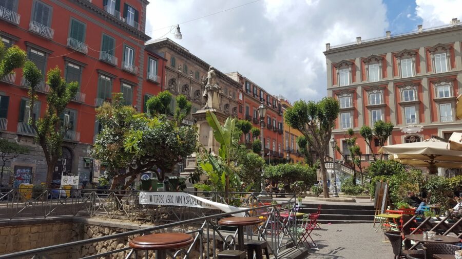City plaza with outdoor cafe, statue, cobblestone square, and colorful European buildings.