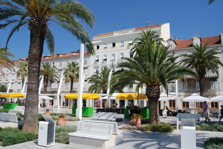 Vibrant Mediterranean plaza with palms, dining, and mixed architecture in coastal city.